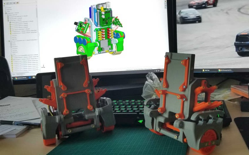 Two 3D-printed Bobble-Bot chassis on a cutting mat, with the CAD model on the monitor behind them